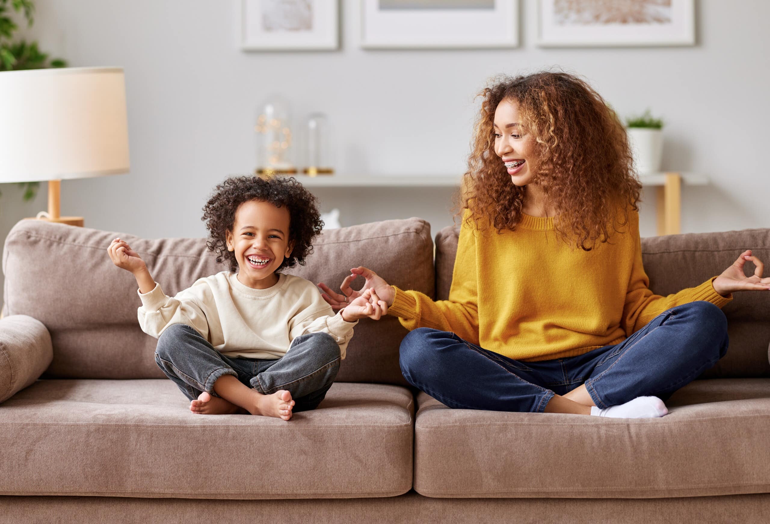 Yoga with kids. Young afro american family mother and little son meditating while sitting in lotus pose on sofa in living room, smiling and enjoying leisure time together at home